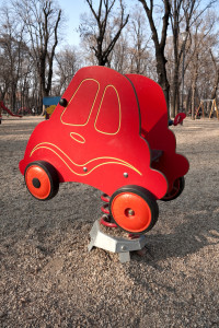 Bright red toy car made from a plastic material resembles the shape of a beetle. It is situated inside a children's playground in an area covered with gravel and sand. It stands on a large spring. In the background there are several tall trees.